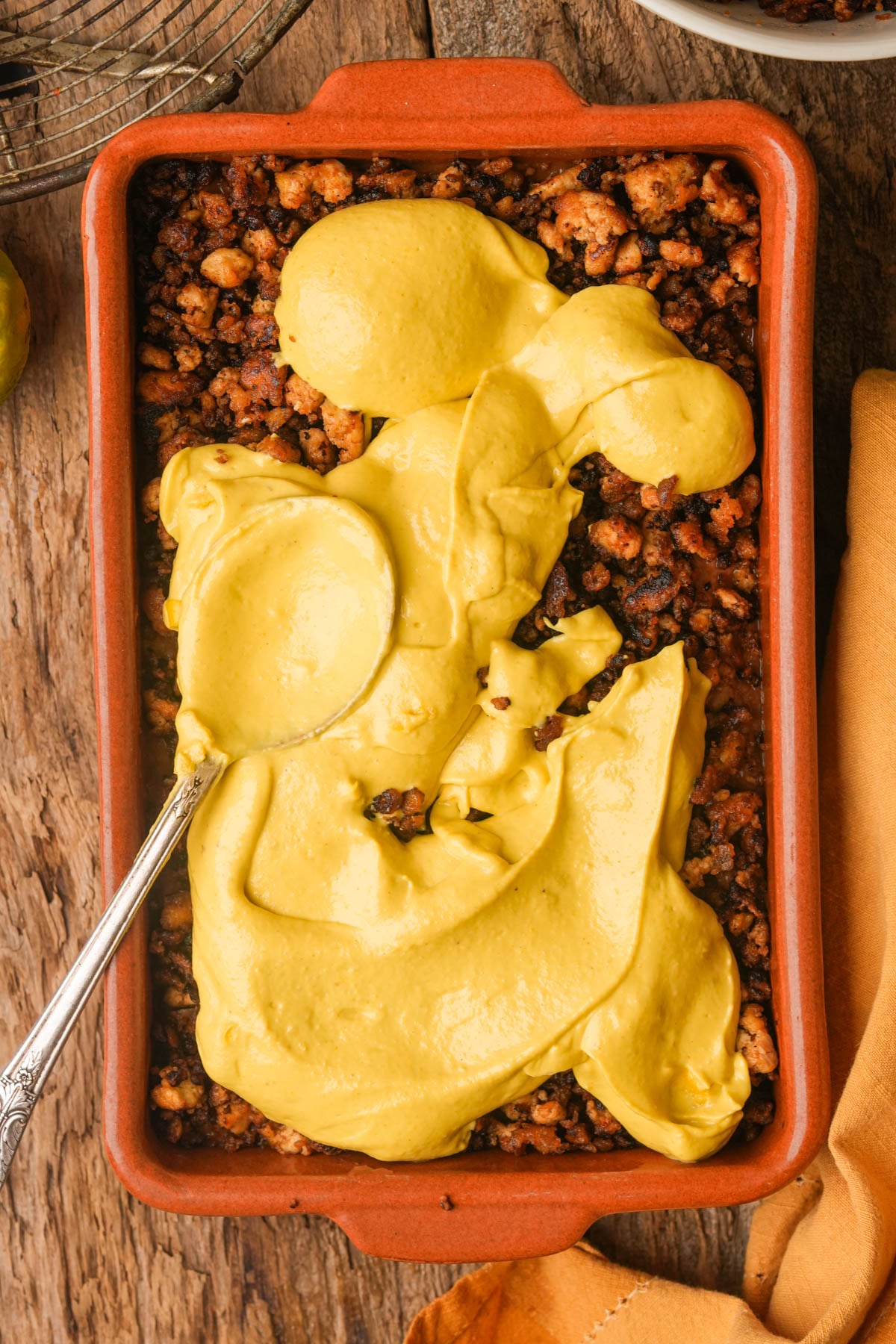 A rectangular baking dish filled with a vegan chorizo topped with thick vegan queso; a spoon rests in the dish, partially spreading the sauce. The dish sits on a rustic wooden surface.