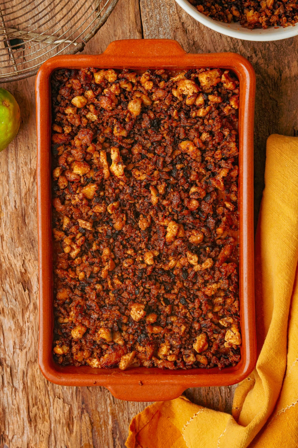 A rectangular baking dish filled with browned, crumbled vegan chorizo sits on a wooden surface next to a yellow napkin and a bowl with more of the mixture.