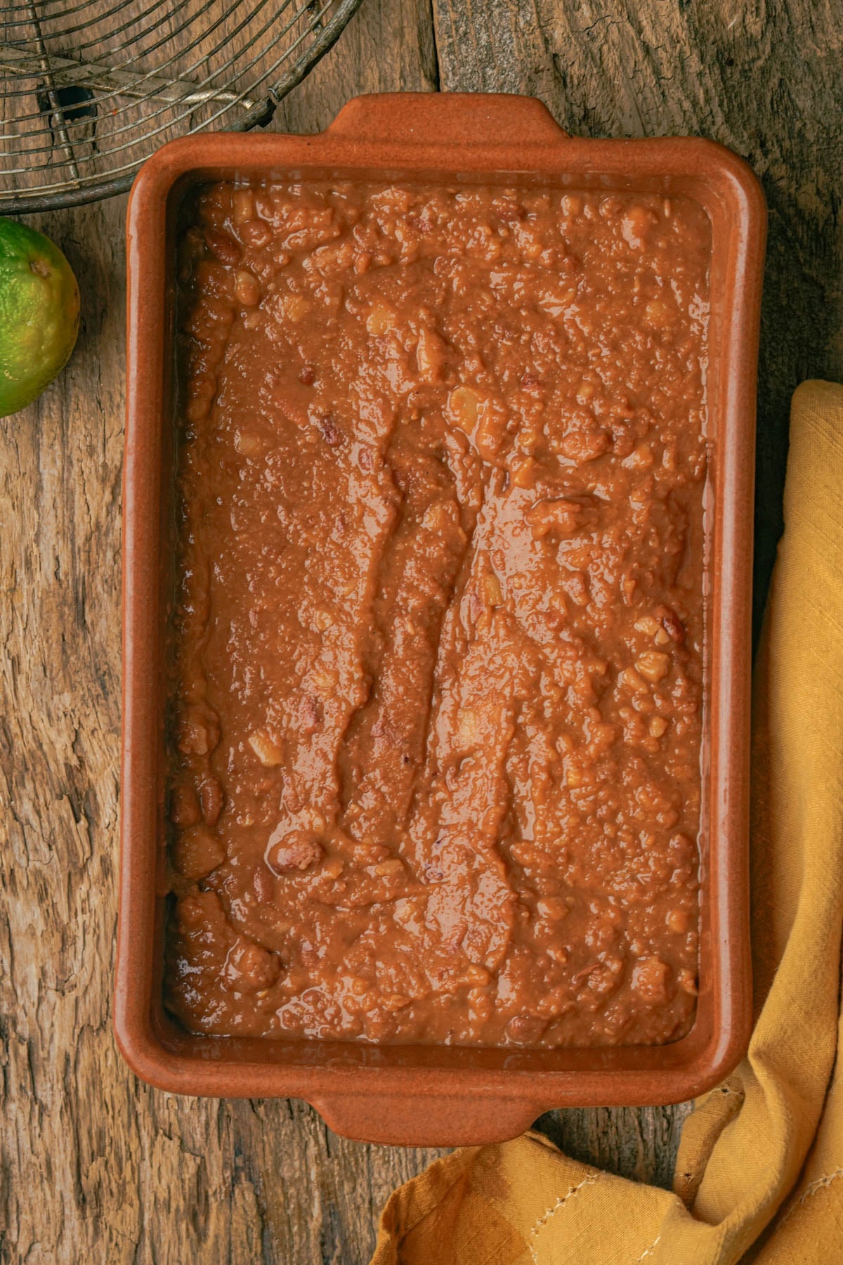 A rectangular brown dish filled with refried beans sits on a rustic wooden table beside a yellow cloth and a lime, with a metal cooling rack partially visible in the corner.