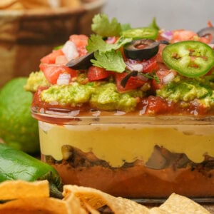 A vegan seven-layer dip in a glass dish, featuring layers of refried beans, cheese, guacamole, salsa, tomatoes, onions, olives, jalapeños, and cilantro, with tortilla chips and limes nearby.