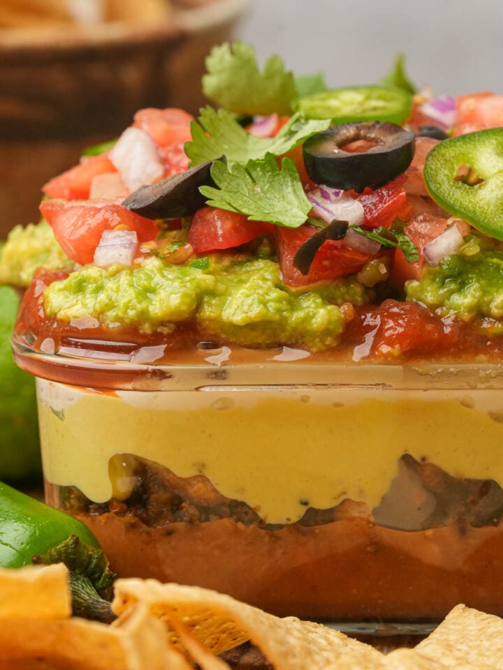 A vegan seven-layer dip in a glass dish, featuring layers of refried beans, cheese, guacamole, salsa, tomatoes, onions, olives, jalapeños, and cilantro, with tortilla chips and limes nearby.
