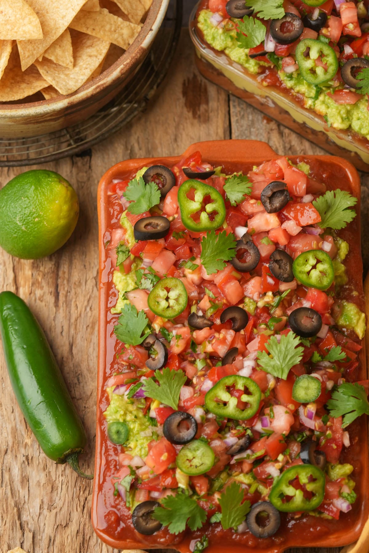 A rectangular dish of layered vegan seven-layer dip topped with chopped tomatoes, black olives, jalapeño slices, cilantro, and onions sits on a wooden table next to a lime, a jalapeño pepper, and a bowl of tortilla chips.