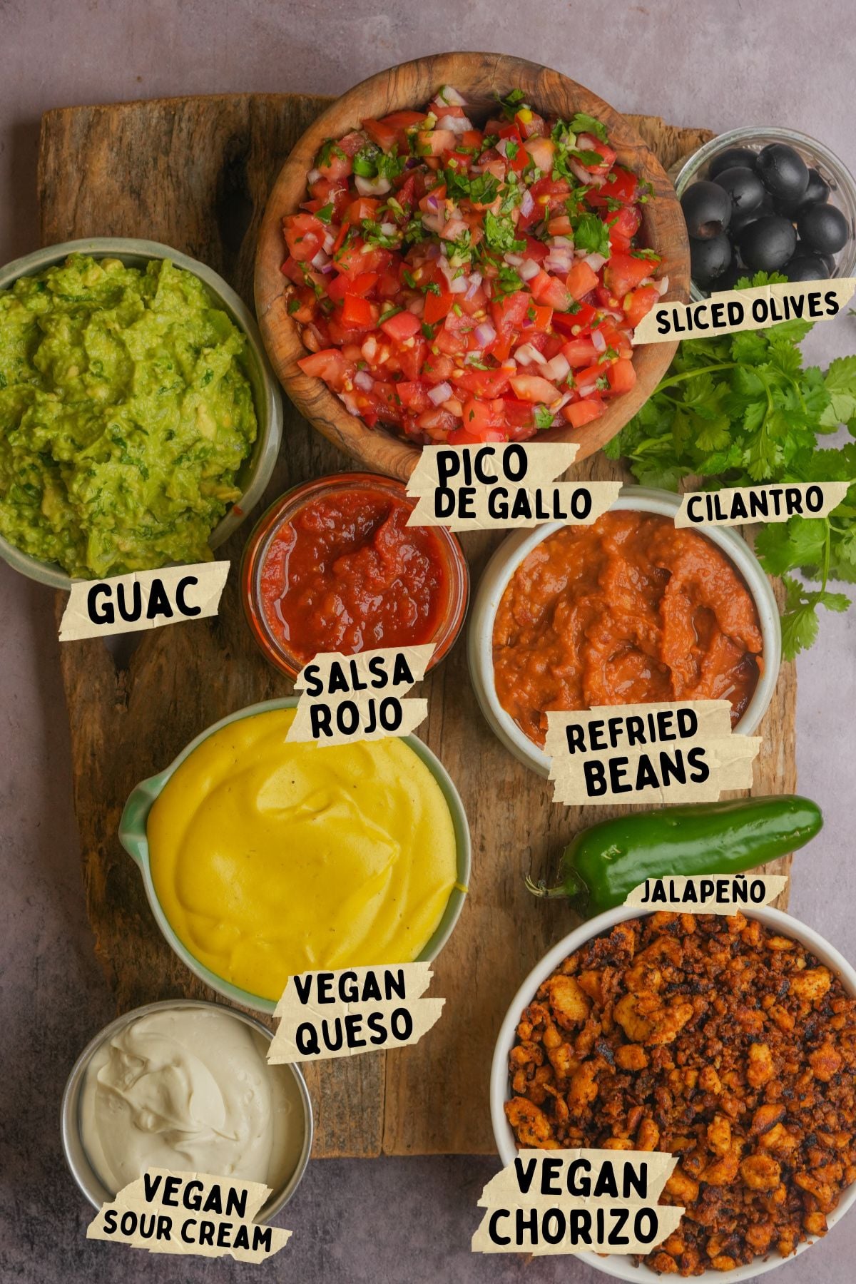 A wooden board topped with bowls of guacamole, pico de gallo, salsa rojo, refried beans, vegan queso, vegan chorizo, vegan sour cream, sliced olives, and fresh cilantro, with a jalapeño pepper on the side.