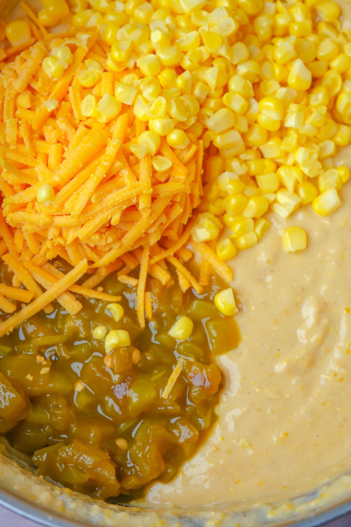 Close-up of a mixing bowl containing shredded vegan cheddar cheese, whole kernel corn, chopped green chiles, and a creamy yellow vegan tamale pie topping batter. The ingredients are not yet mixed together.