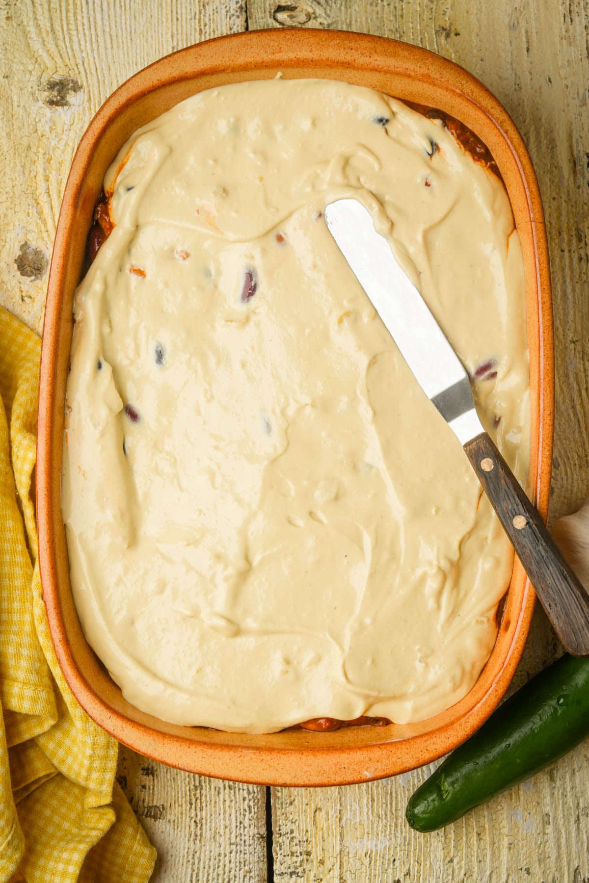 A layer of vegan queso is spread across a pan of vegan tamale pie. A yellow checkered cloth and a green jalapeño are beside the dish on a rustic wooden surface.