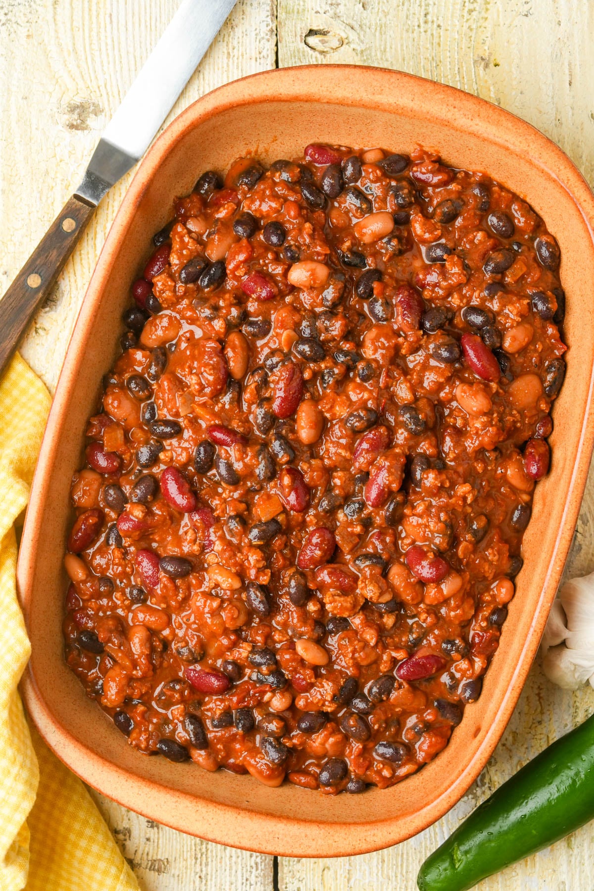 A baking dish filled with chili made of beans, ground seitan, and tomato sauce sits on a wooden surface. A knife, yellow cloth, garlic, and a green chili pepper are nearby.