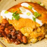 A serving of vegan tamale pie topped with sour cream, chopped green onions, and cilantro, served with a side of seasoned black and pinto beans on a plate.
