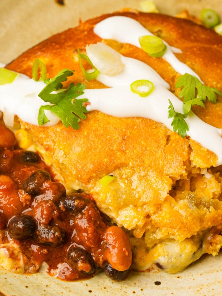 A serving of vegan tamale pie topped with sour cream, chopped green onions, and cilantro, served with a side of seasoned black and pinto beans on a plate.