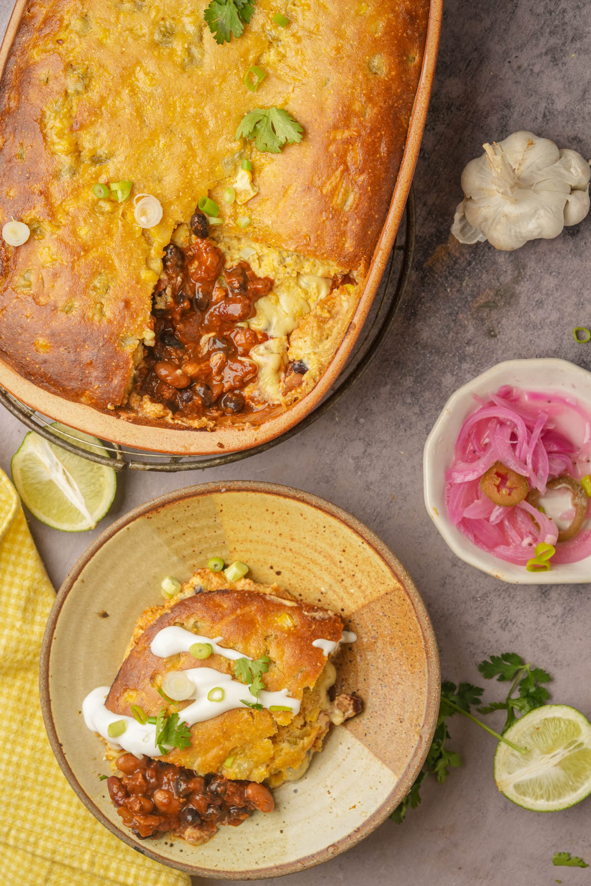 A casserole dish filled with a golden baked vegan tamale pie, partially served onto a bowl. The bowl contains a slice topped with sour cream, cilantro, and green onions. Nearby are pickled onions, garlic, lime wedges, and a yellow napkin.