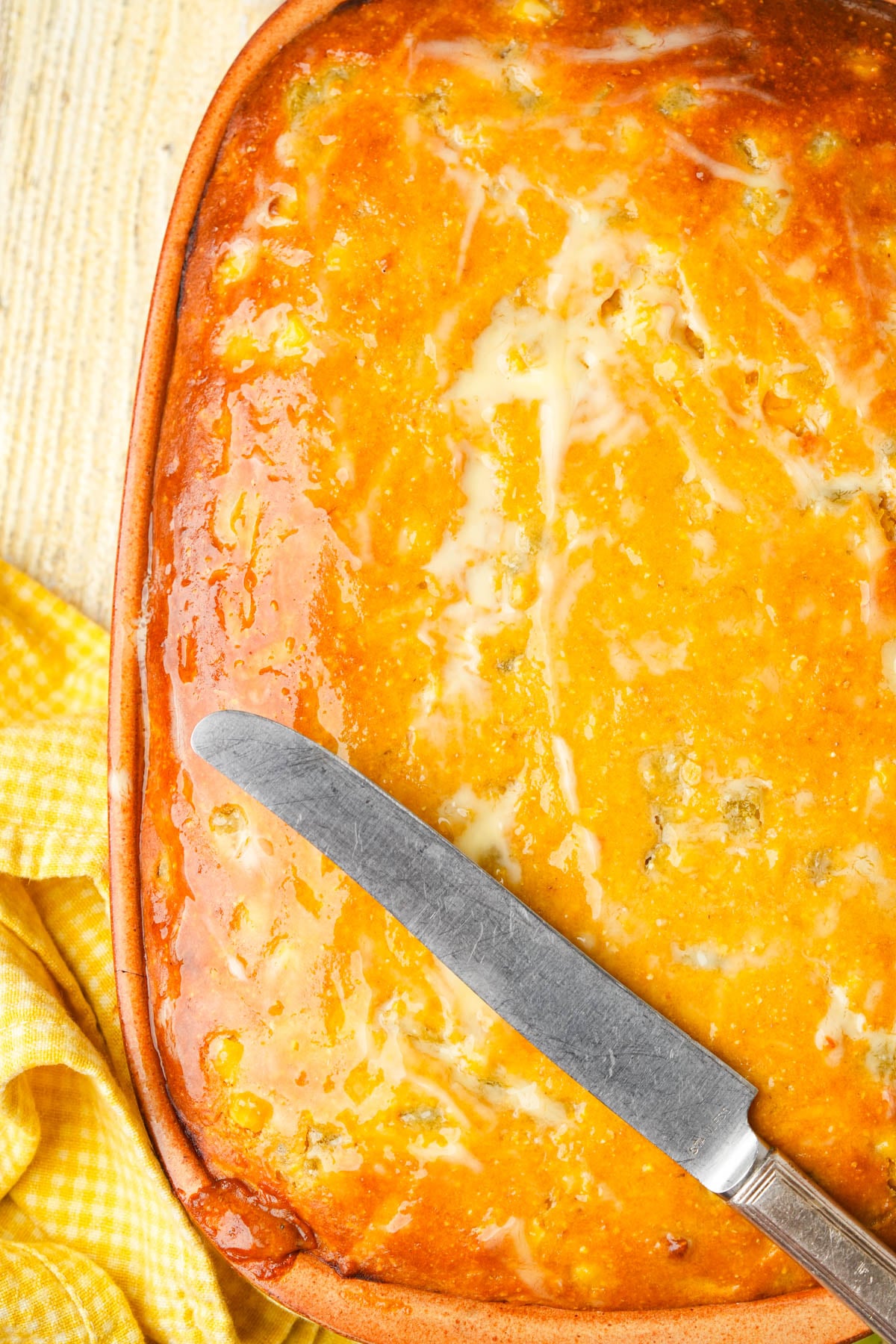 A baked vegan tamale pie with a golden, glossy top layer, coated in melted vegan butter a knife resting on it. A yellow checkered cloth sits beside the dish on a wooden surface.