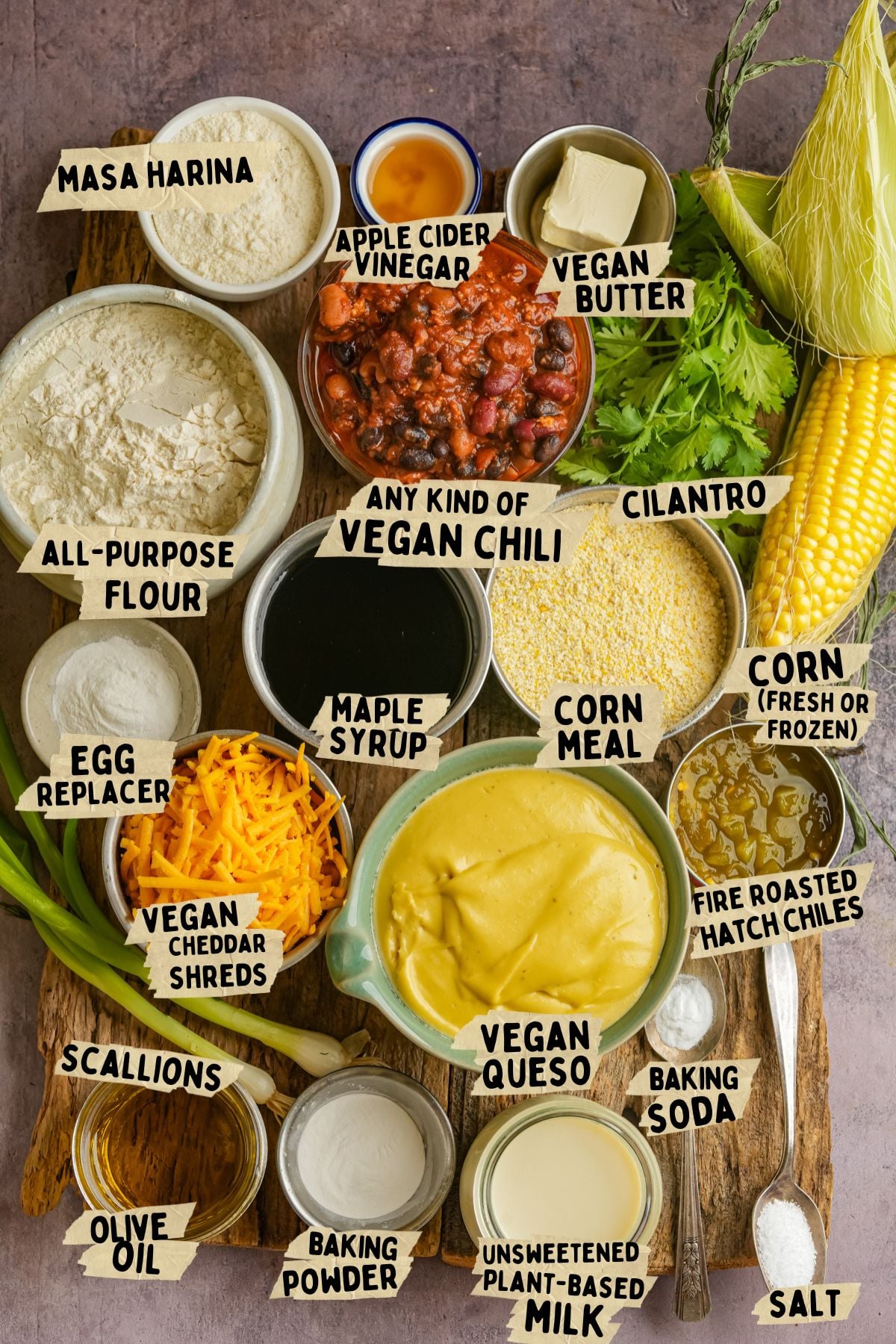 A wooden tray holds labeled ingredients for a vegan tamale pie recipe: masa harina, all-purpose flour, apple cider vinegar, vegan butter, vegan chili, cilantro, corn, cornmeal, scallions, olive oil, egg replacer, vegan cheddar shreds, maple syrup, fire roasted Hatch chiles, baking powder, unsweetened plant-based milk, vegan queso, baking soda, and salt.