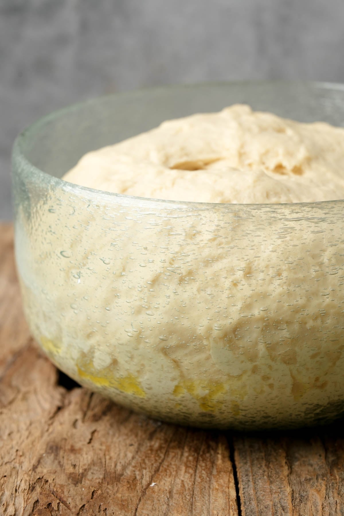 A glass bowl filled with risen bazlama dough sits on a rustic wooden surface, showing bubbles and an airy texture.