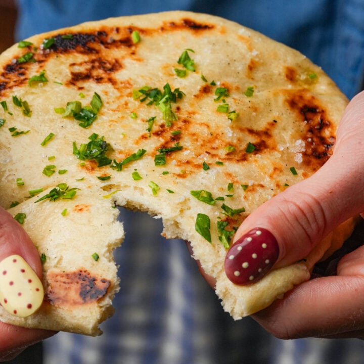A person with polka-dot nail art holds a round, grilled bazlama topped with chopped herbs, showing a large bite taken out of it.