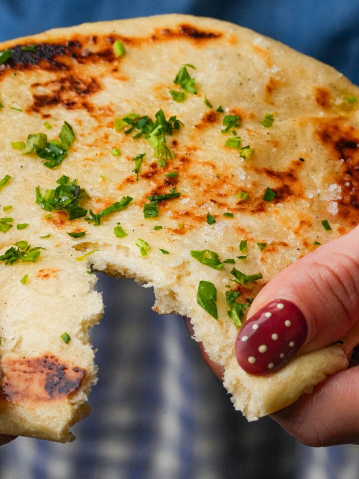 A person with polka-dot nail art holds a round, grilled bazlama topped with chopped herbs, showing a large bite taken out of it.