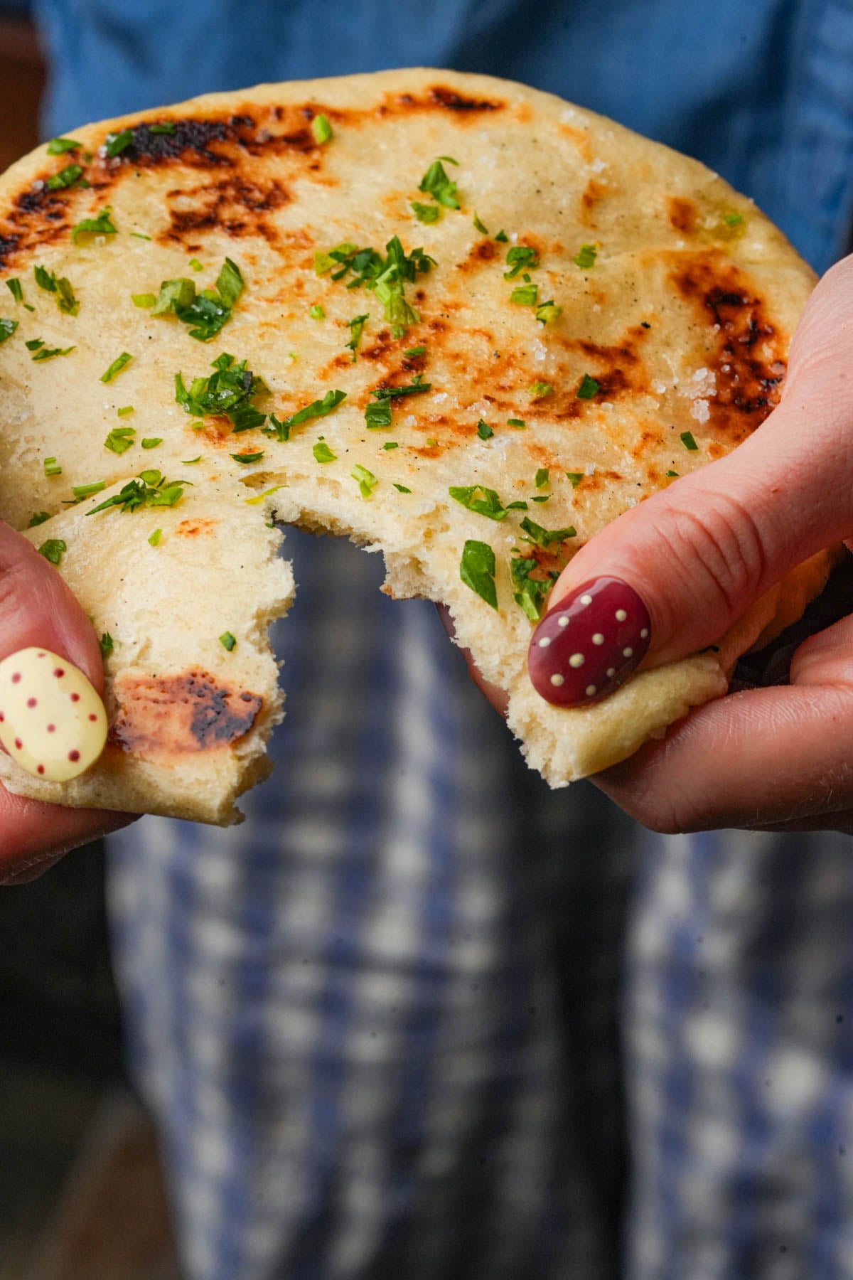 A person with painted nails holds a piece of bazlama with herbs sprinkled on top. The bread has a bite taken out. The person is wearing a blue shirt and plaid pants.