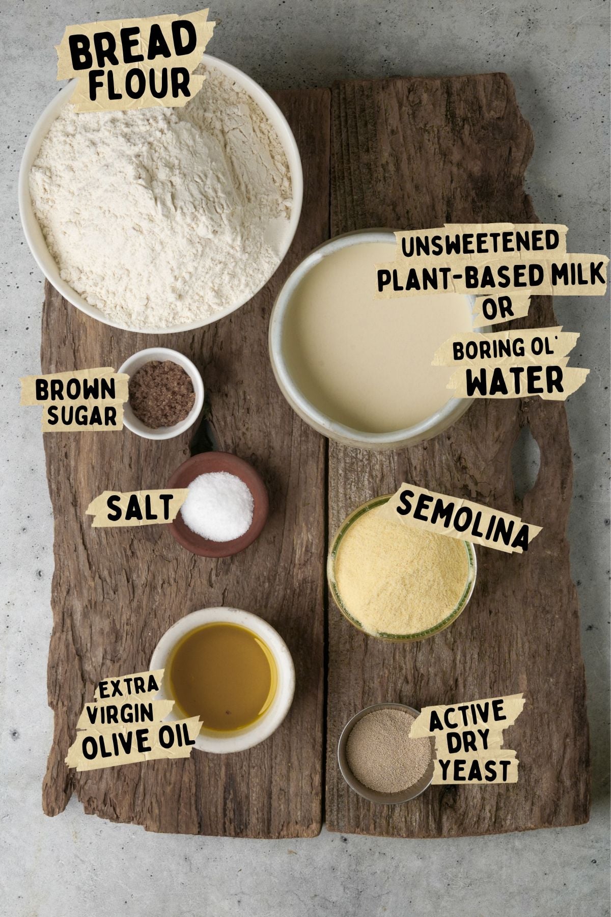 Ingredients for Bazlama bread: bowl of bread flour, plant-based milk (or water), brown sugar, salt, semolina, extra virgin olive oil, and active dry yeast, all labeled and arranged on a rustic wooden board.