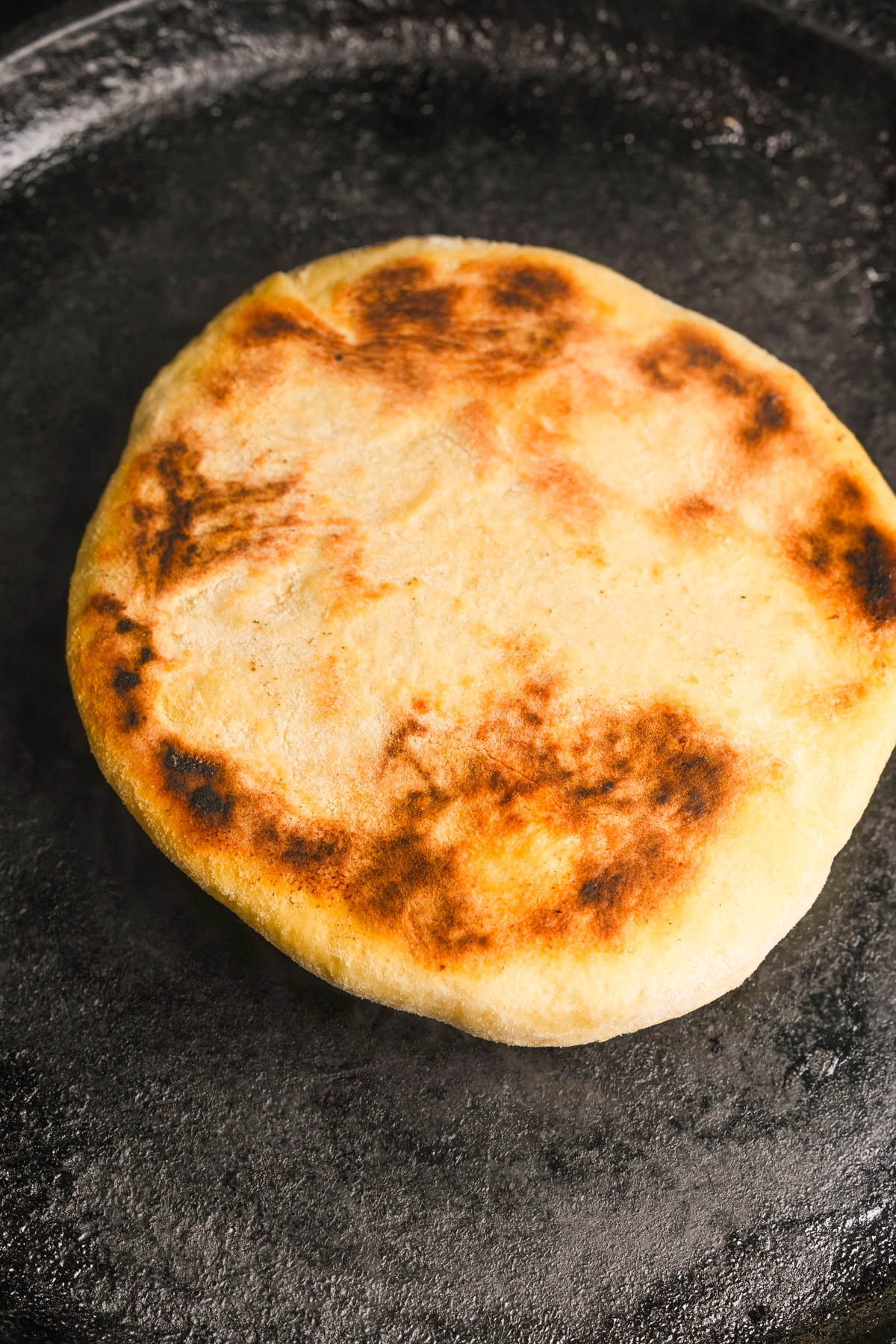 A golden-brown bazlama with charred spots sits on a dark, textured surface, possibly a skillet or griddle.