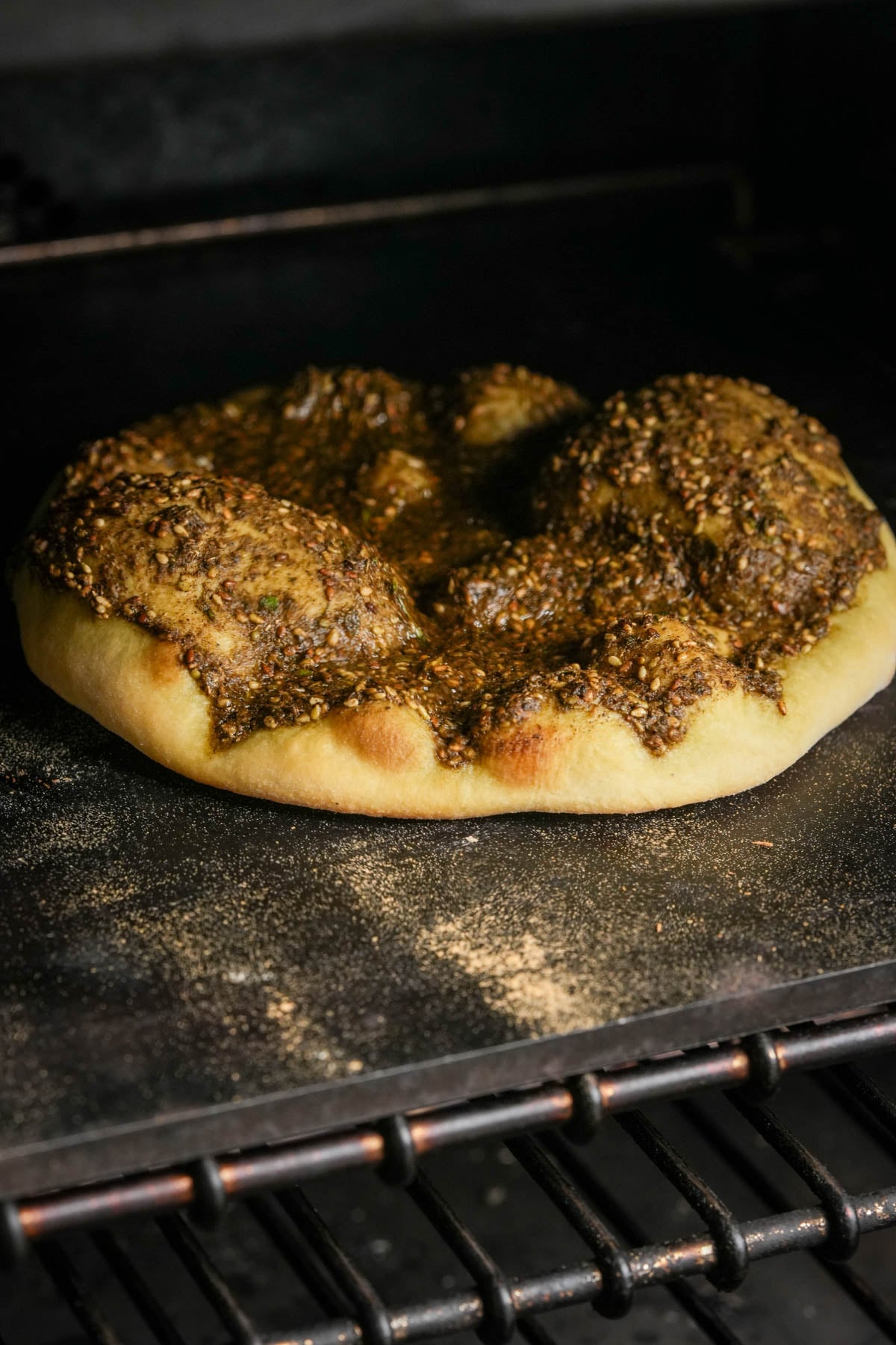 Manakeesh flatbread topped with za'atar seasoning baking on a pizza stone in an oven, with some flour scattered on the stone and oven racks visible in the foreground.