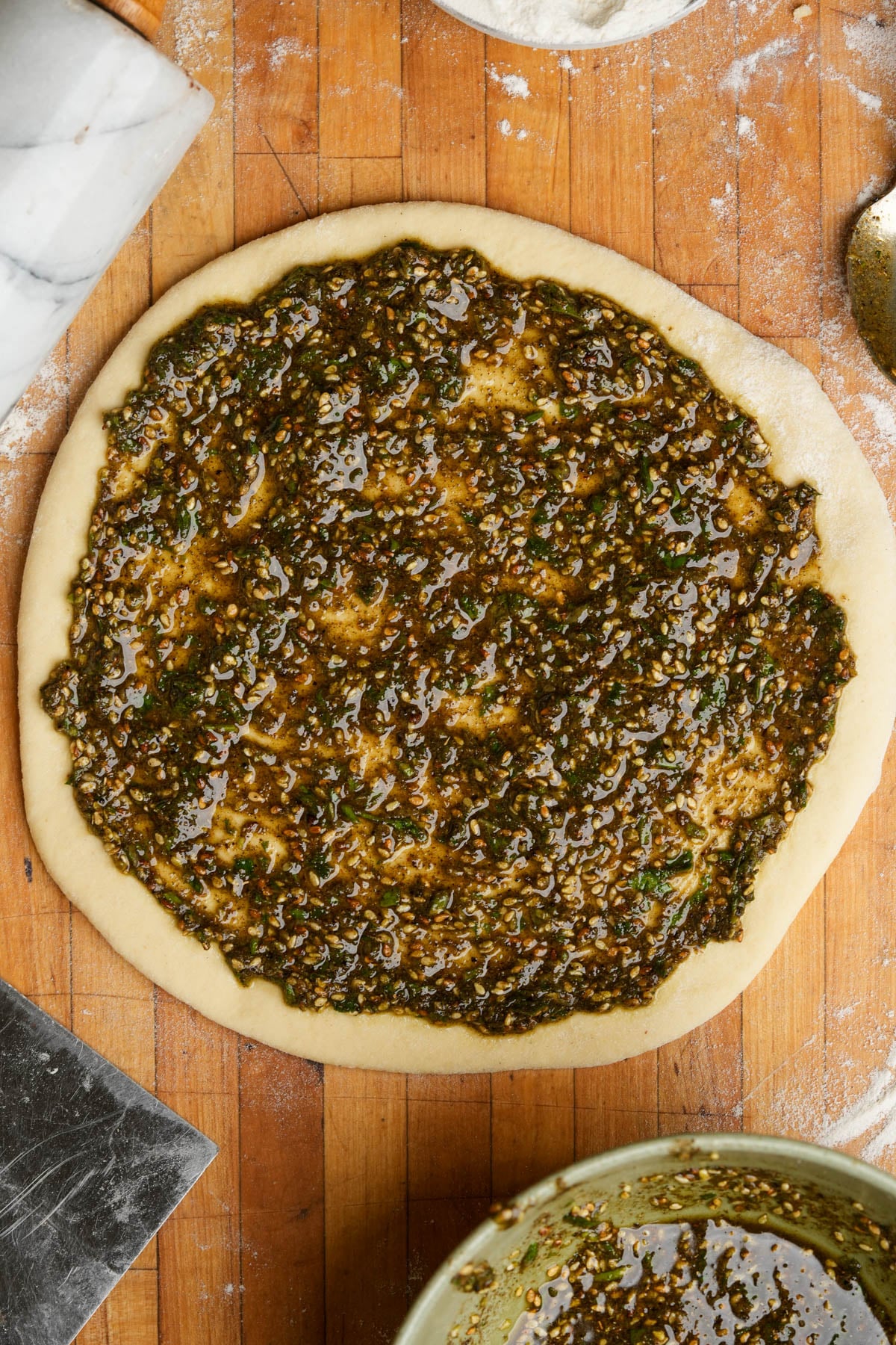 Round manakeesh dough topped with a thick, greenish-brown za'atar and herb mixture sits on a floured wooden surface. Kitchen tools and a bowl with more pesto are nearby.
