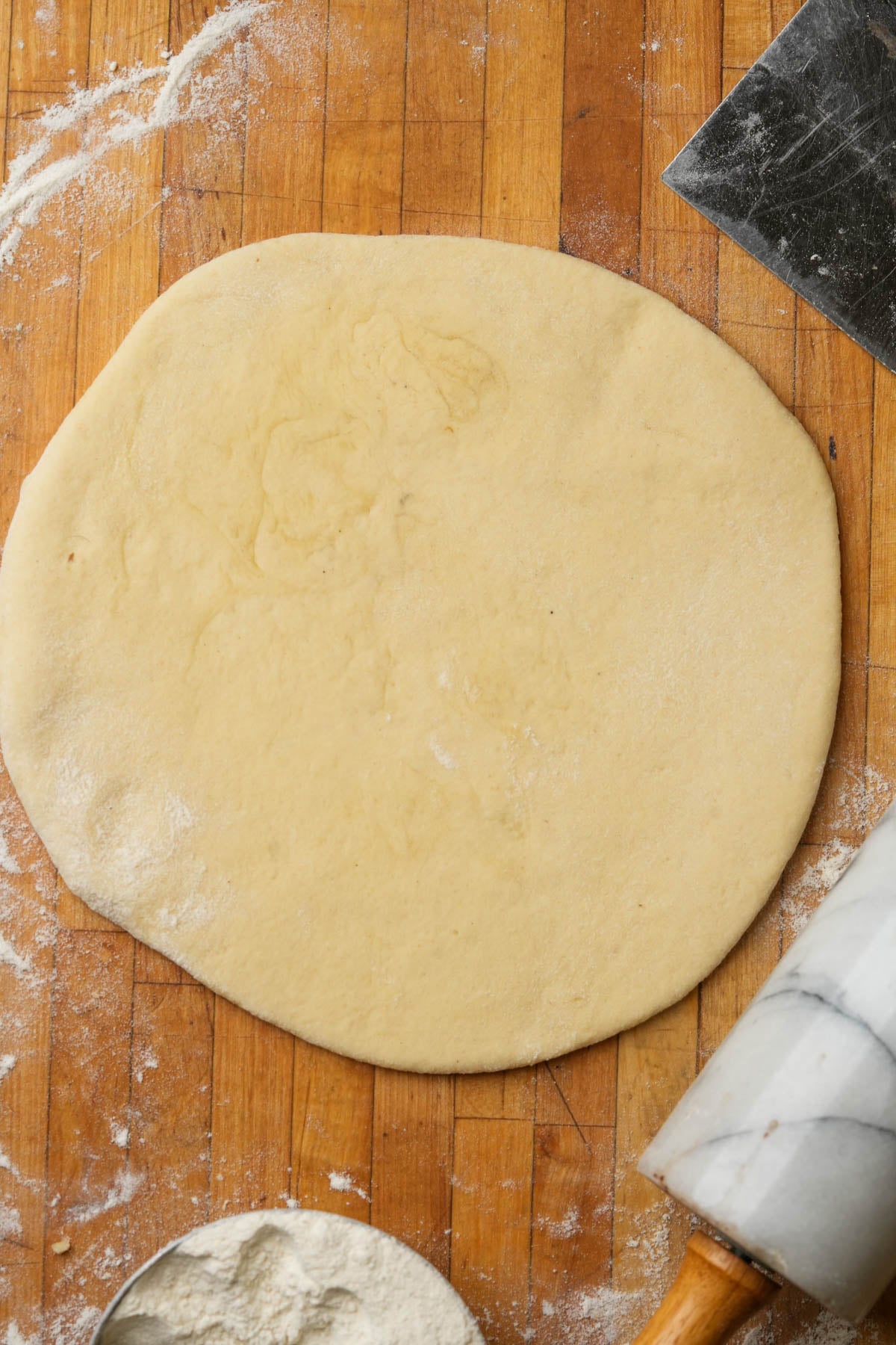 A round sheet of dough on a floured wooden surface, with a marble rolling pin, a metal scraper, and a bowl of flour nearby.