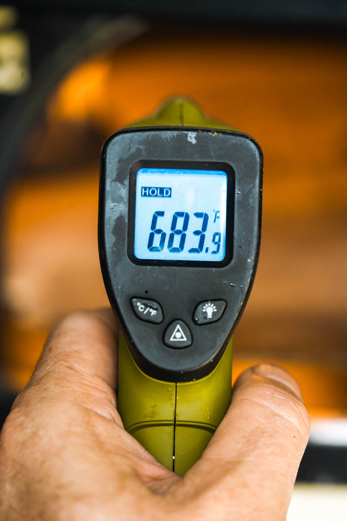 A hand holds a green infrared thermometer displaying a temperature of 683.9°F on its screen, with a blurred orange background.