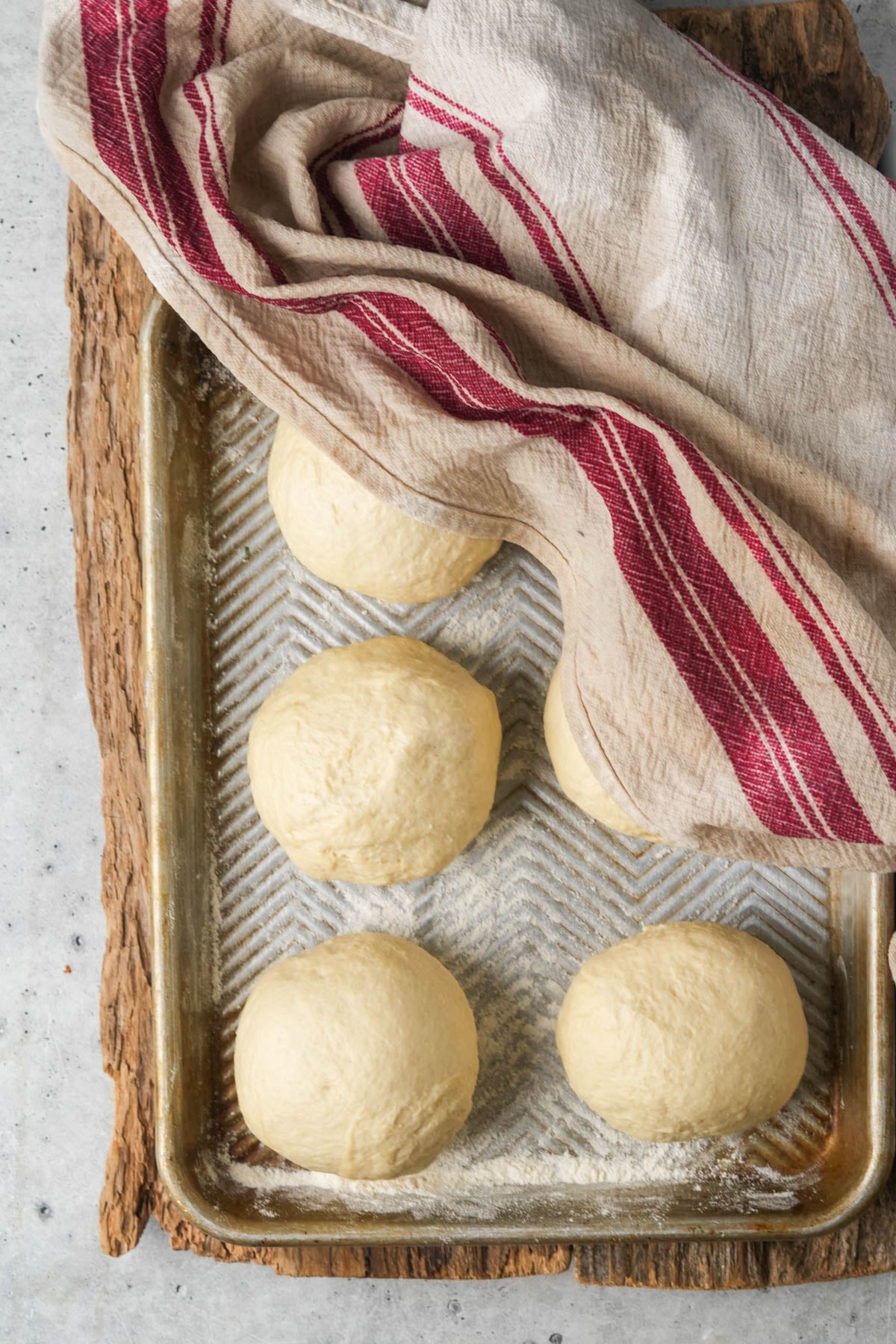 Six Manakeesh dough balls rest on a floured baking tray, partially covered by a beige cloth with red stripes. The tray sits on a rustic wooden surface.