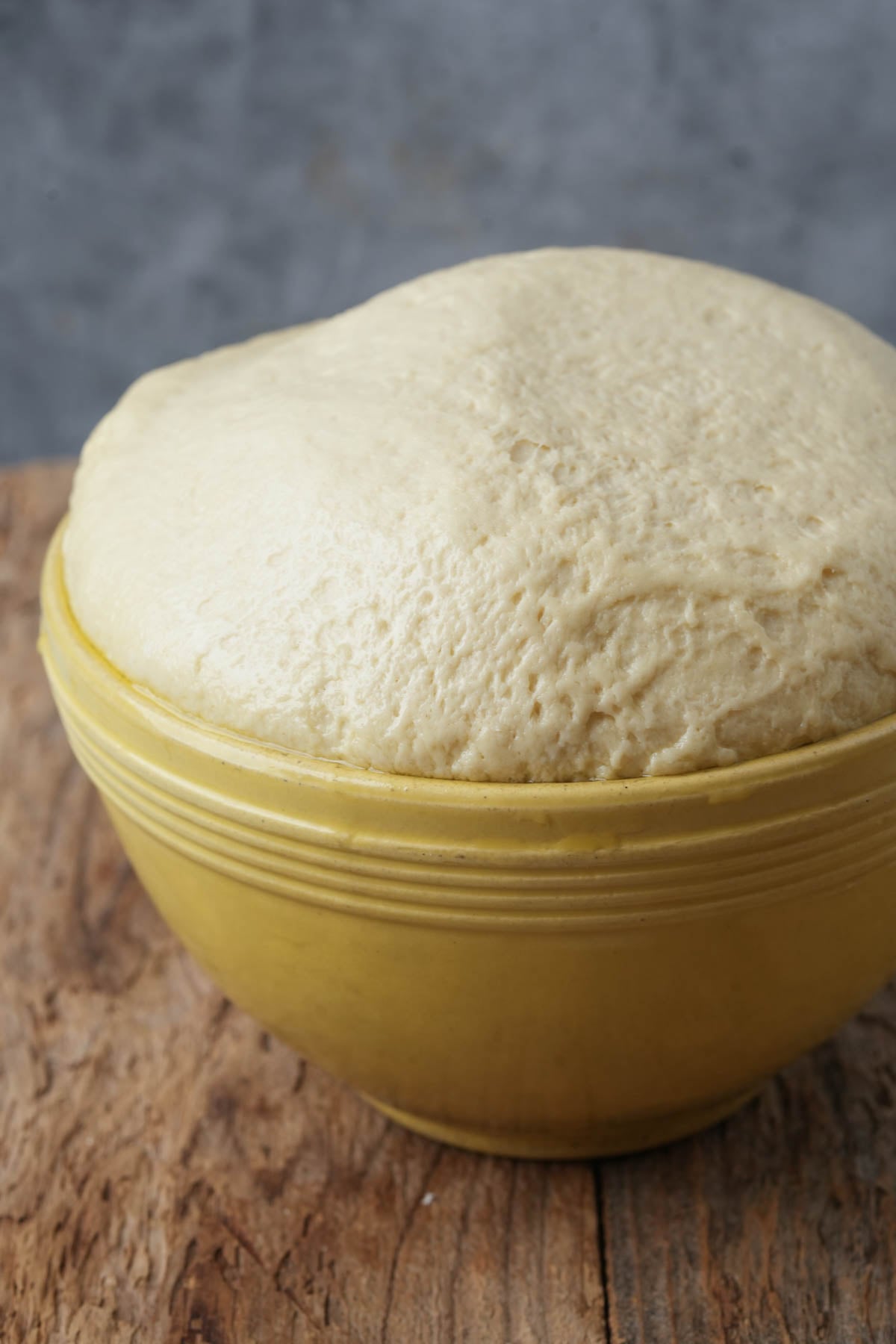 A yellow bowl filled with risen Manakeesh dough sits on a wooden surface against a gray background. The dough is puffed up, overflowing slightly above the bowl's rim.