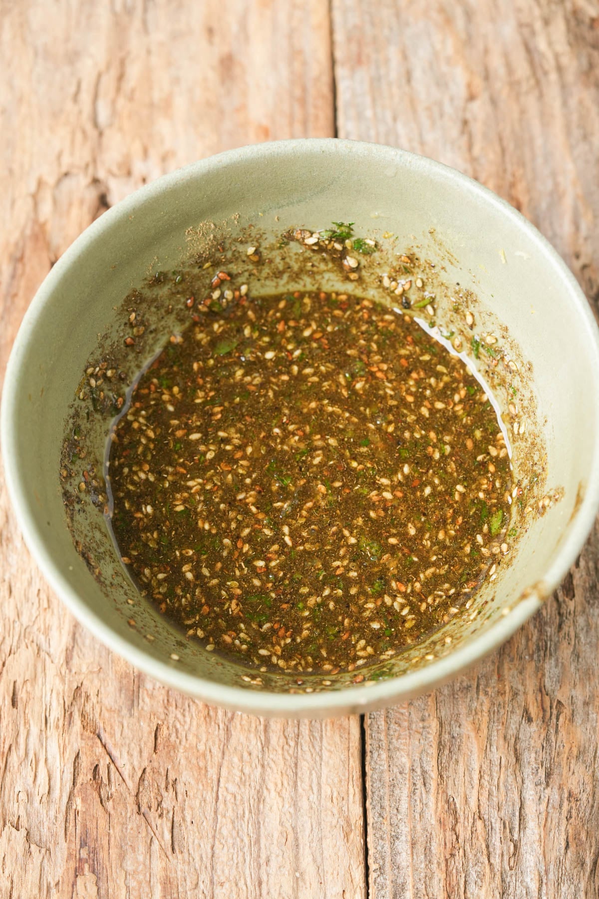 A green bowl filled with za'atar Manakeesh topping sits on a rustic wooden surface.