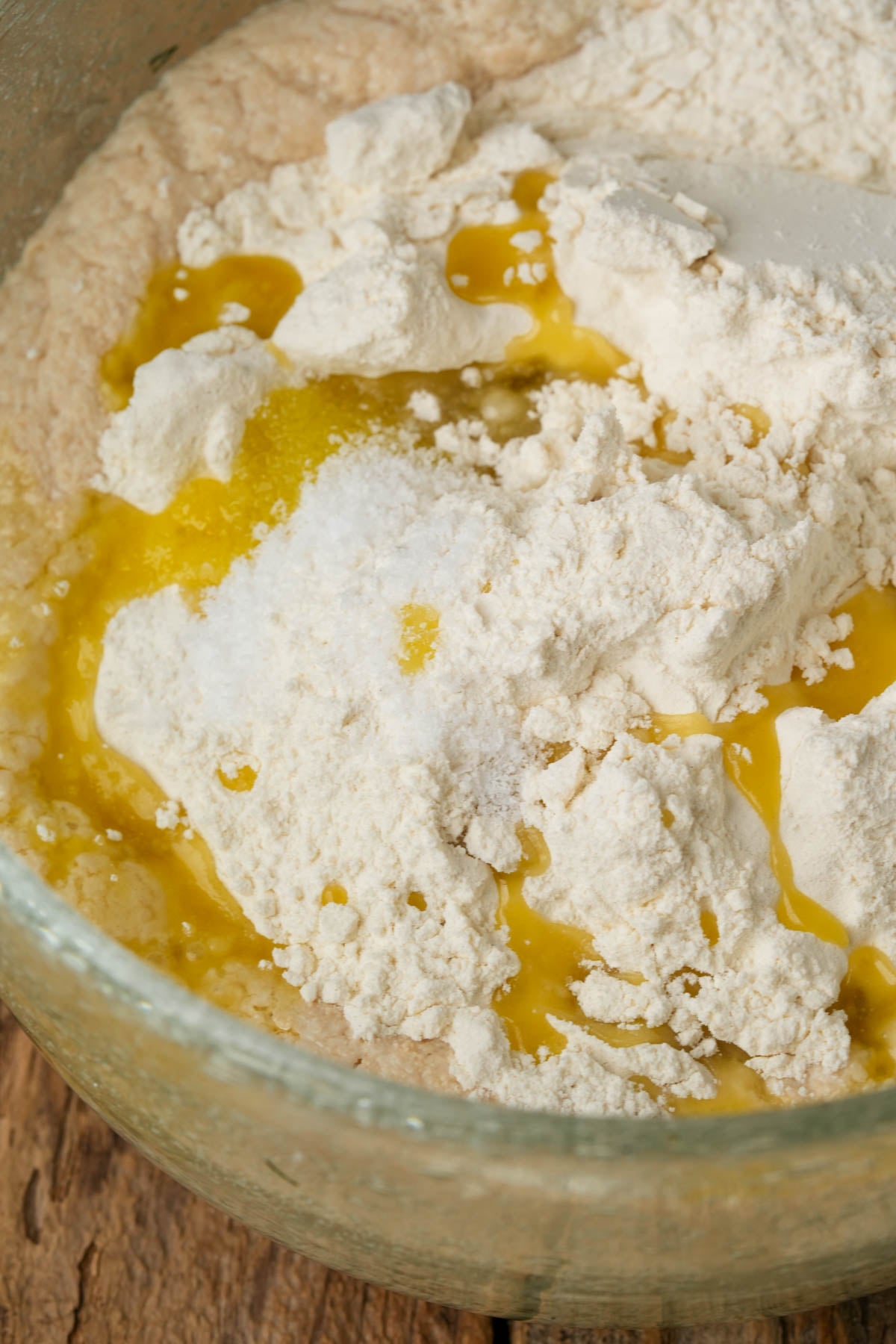 A glass bowl containing flour, melted butter, and other ingredients being mixed for baking, on a wooden surface.