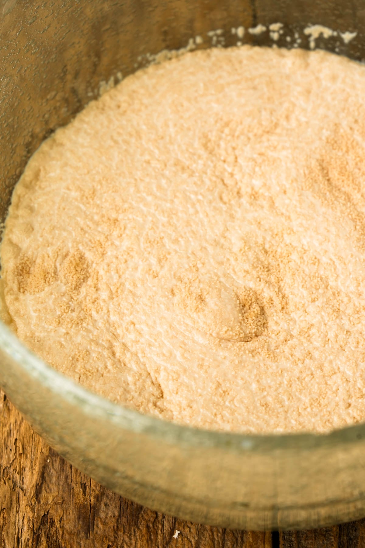 A glass bowl containing a frothy, bubbly mixture of activated yeast, set on a rustic wooden surface.