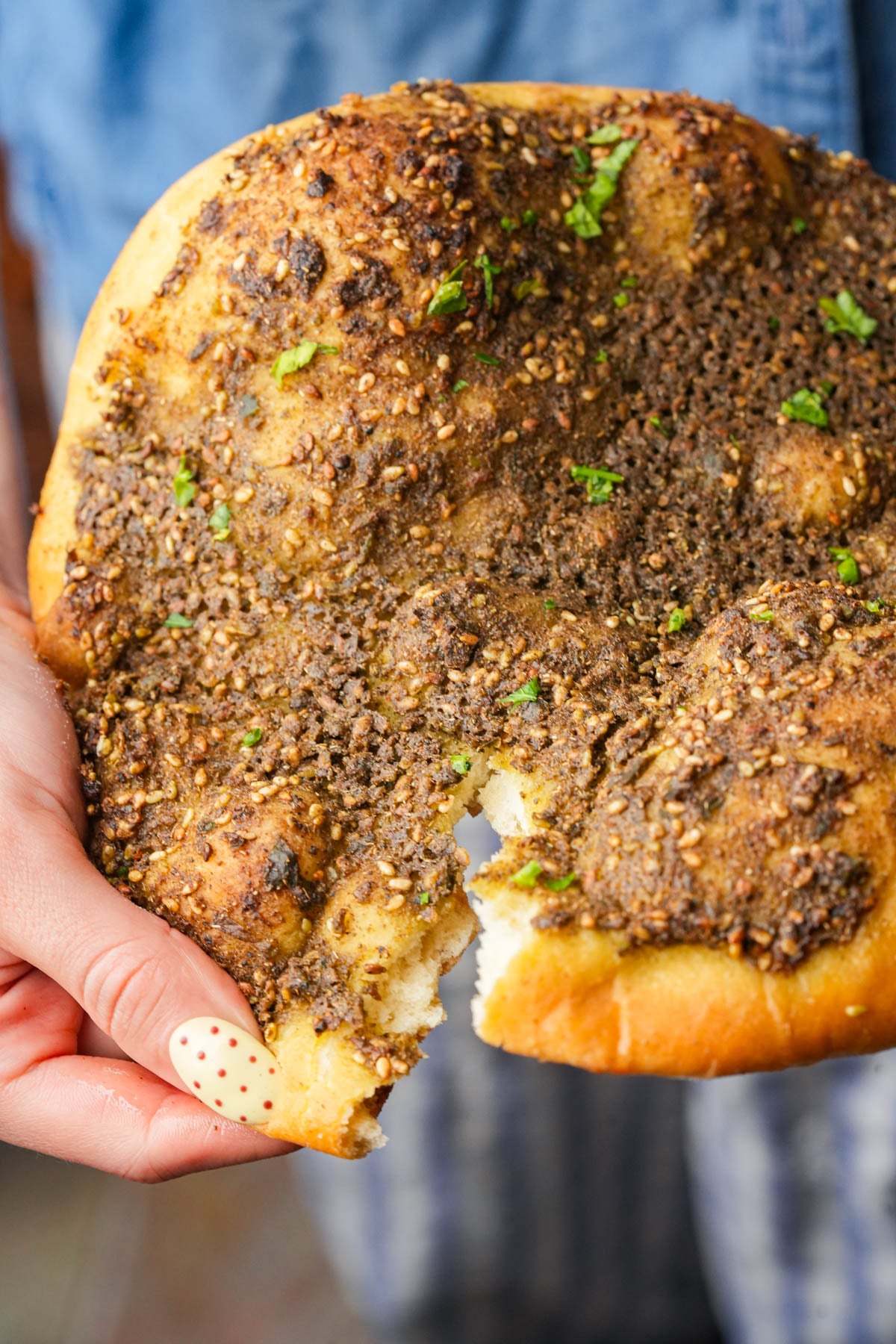 A hand holding a piece of Manakeesh topped with za'atar seasoning, with a chunk torn off. The bread is golden brown and garnished with bits of green herbs.