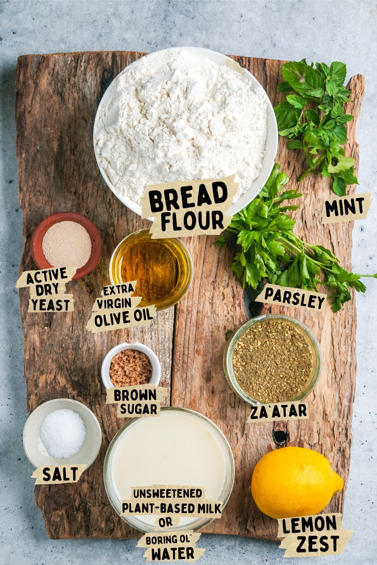A wooden board with labeled bowls of ingredients for a za'atar manakeesh recipe including bread flour, active dry yeast, brown sugar, salt, za'atar, extra virgin olive oil, plant-based milk, parsley, mint, and a lemon with zest.