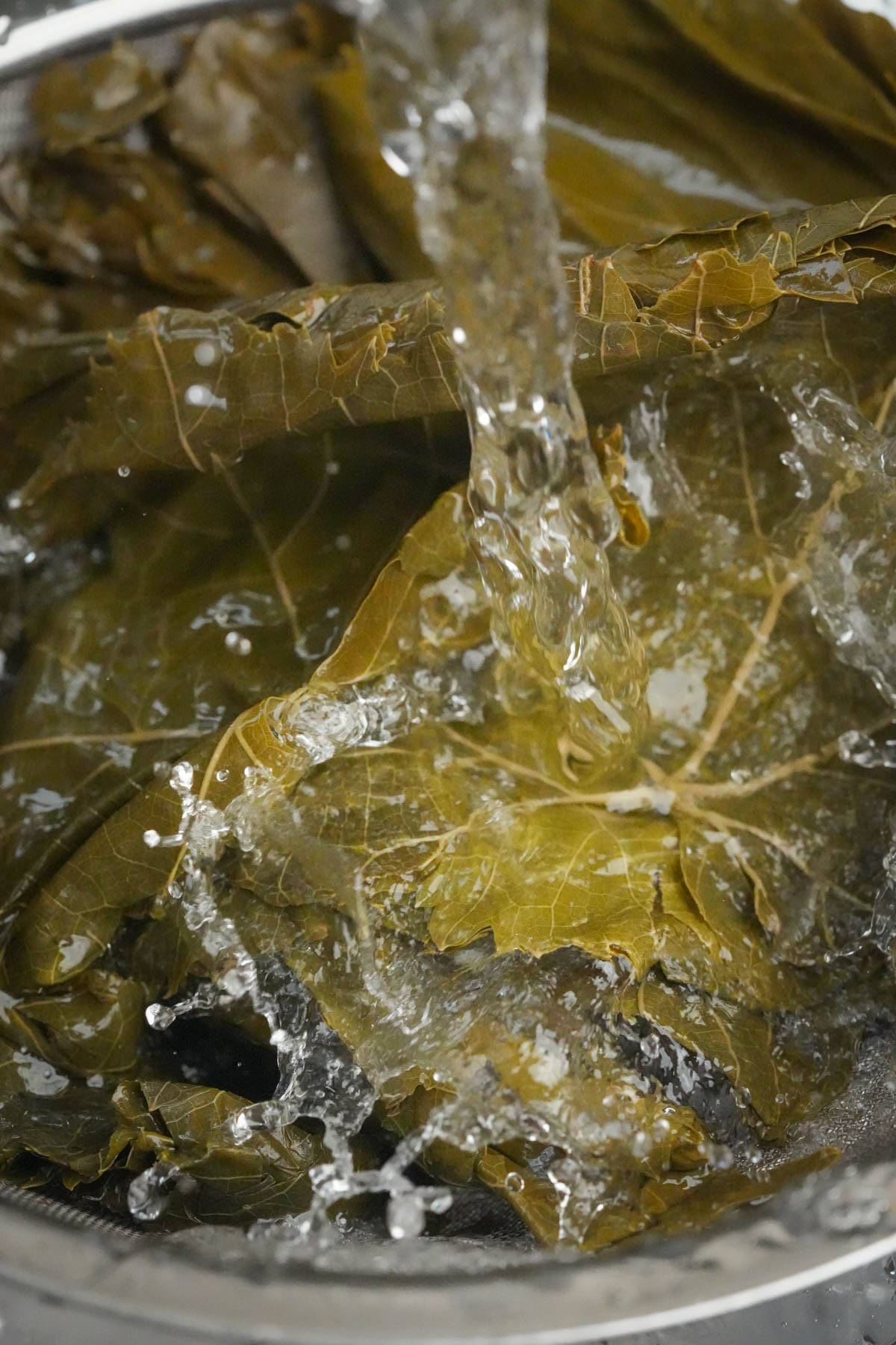 Water is pouring over a pile of grape leaves in a bowl, washing them thoroughly. The leaves are dark green and the water splashes around them.