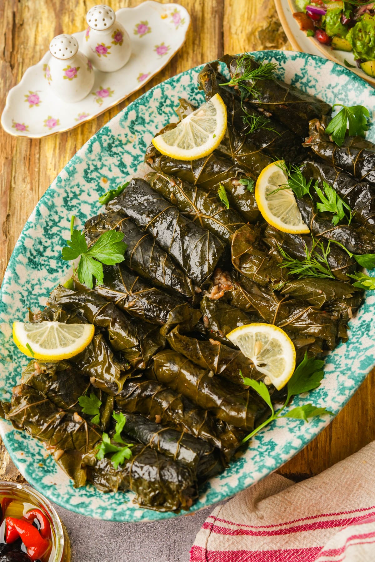 Stuffed yalanji garnished with lemon slices and fresh herbs are arranged on a blue-green oval platter. A floral salt and pepper set, salad, and a striped napkin are visible in the background.