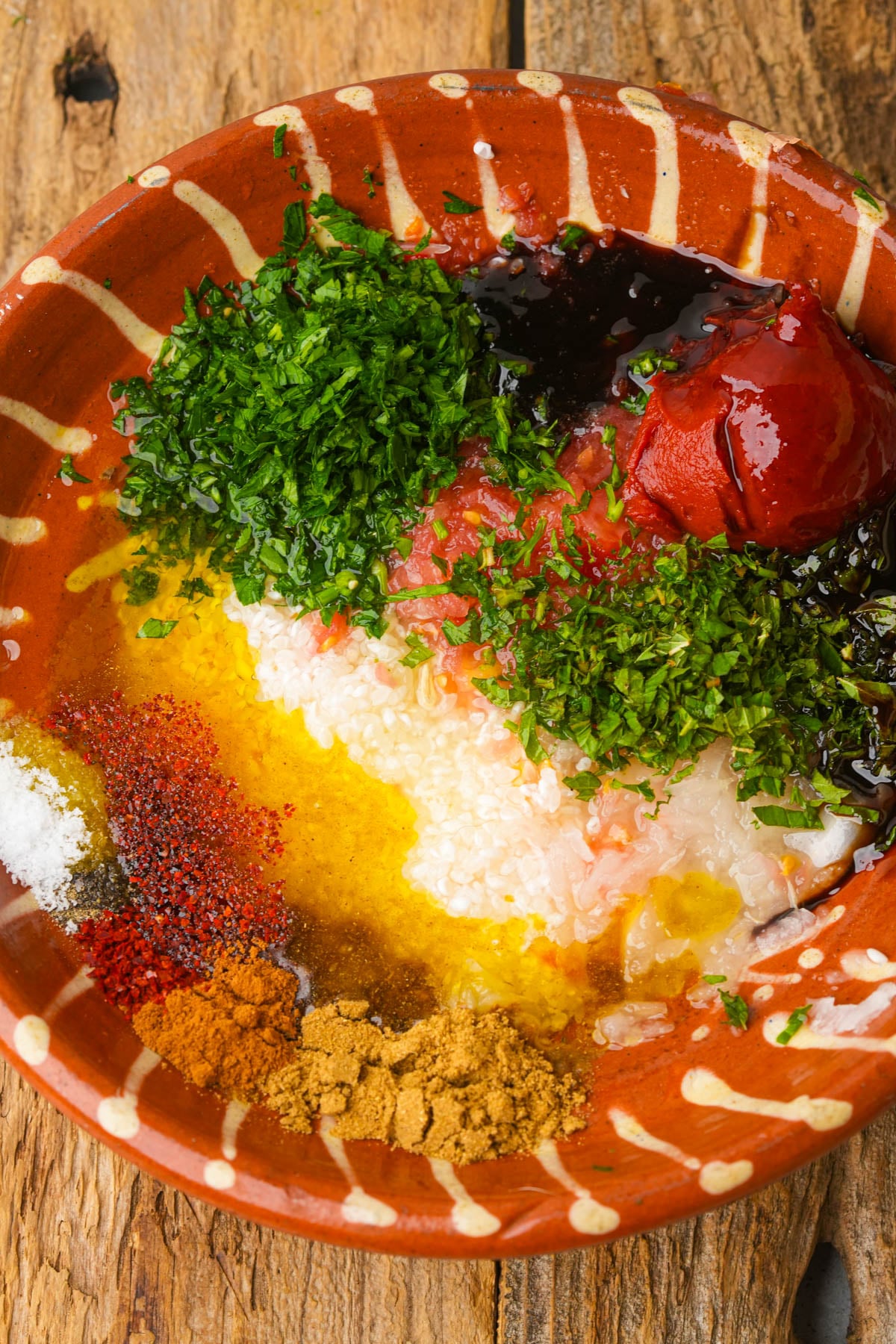 A rustic bowl with chopped herbs, tomato paste, olive oil, spices, minced garlic, and soy sauce on a wooden surface, ready to be mixed fas yalanji filling.