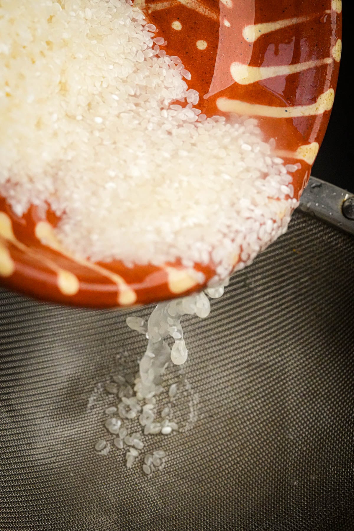 Washed white rice is being poured from a red and yellow patterned bowl into a metal strainer.