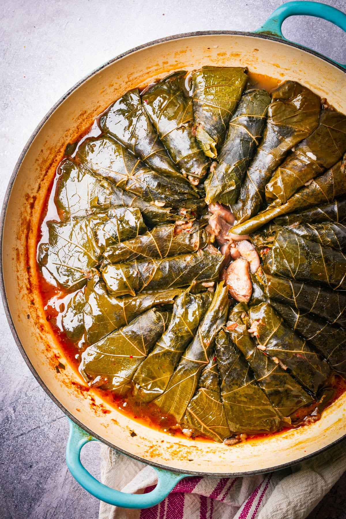 A round pot filled with neatly arranged cooked yalanji, viewed from above. The pot has teal handles and sits on a striped towel.