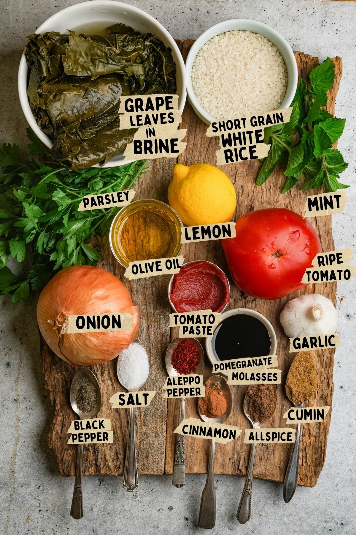 A wooden board with labeled ingredients for a yalanji recipe: grape leaves, white rice, parsley, lemon, mint, tomato, onion, olive oil, tomato paste, pomegranate molasses, garlic, salt, Aleppo pepper, black pepper, cinnamon, allspice, cumin.