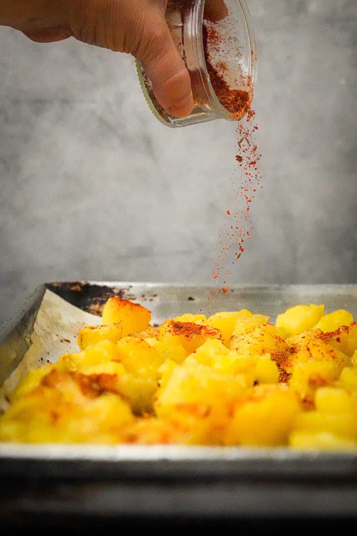 A hand sprinkles red spice from a small jar onto diced yellow potatoes on a baking sheet lined with parchment paper.