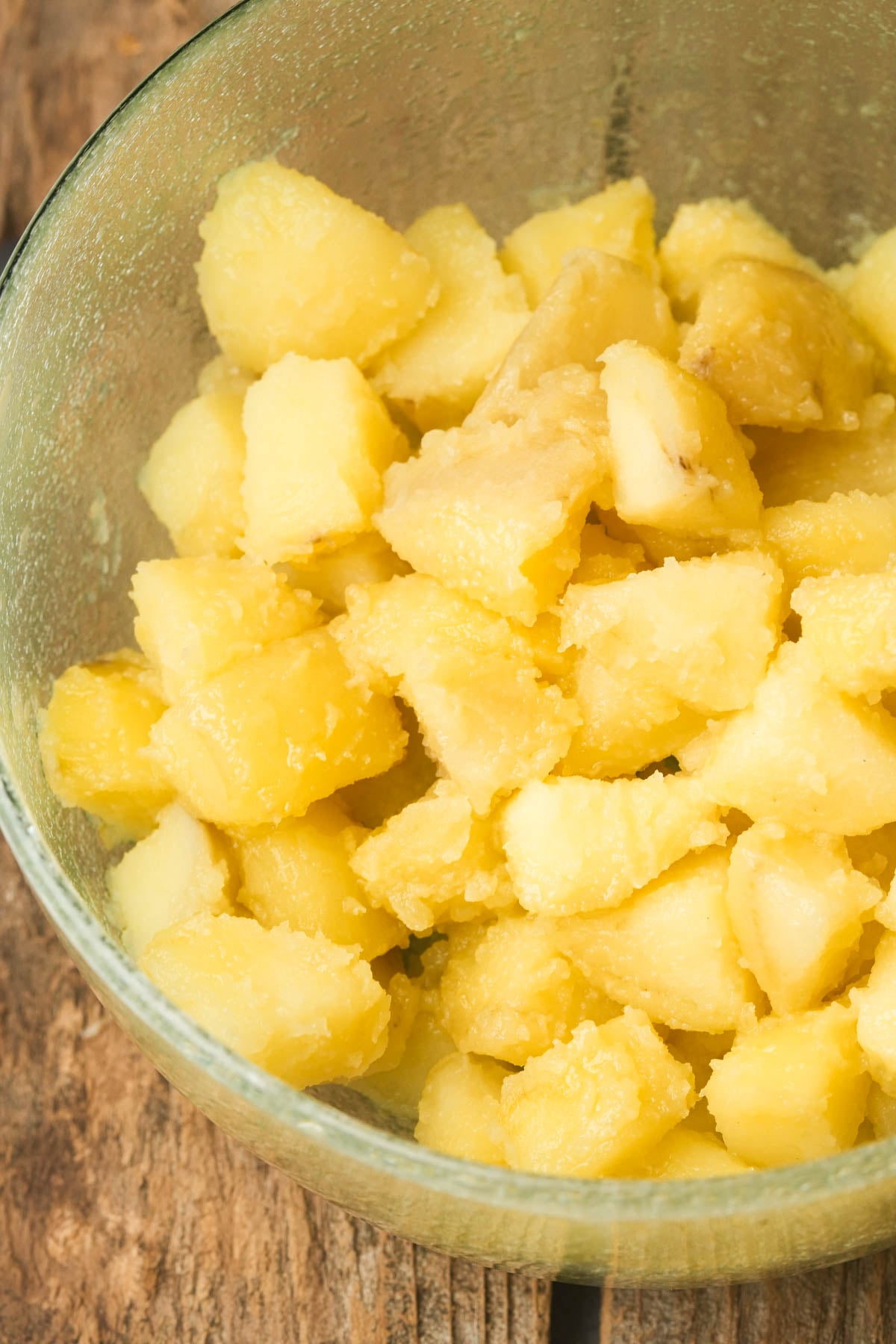 A clear glass bowl filled with beat up par-cooked potatoes sits on a rustic wooden surface.