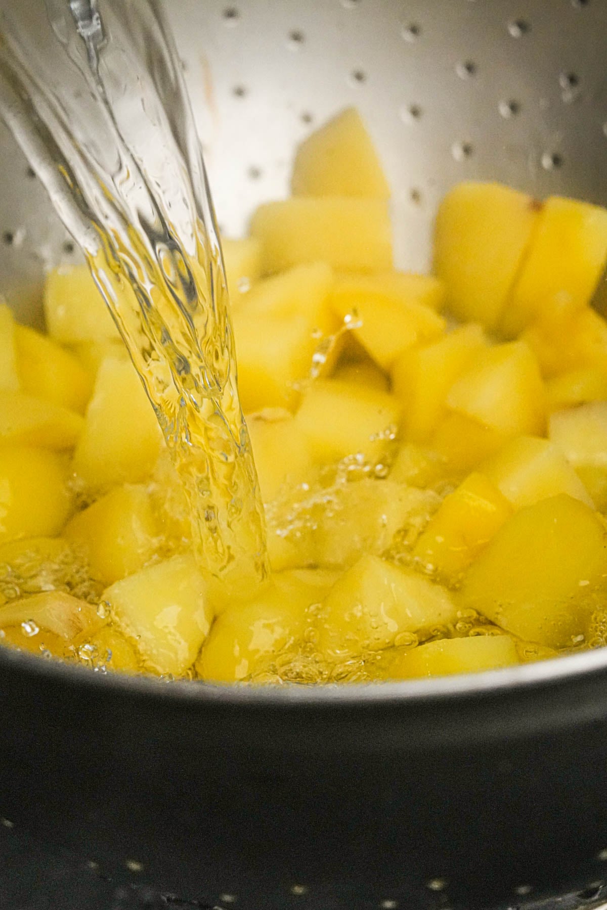 Water being poured over cubed potatoes in a metal colander, rinsing them.