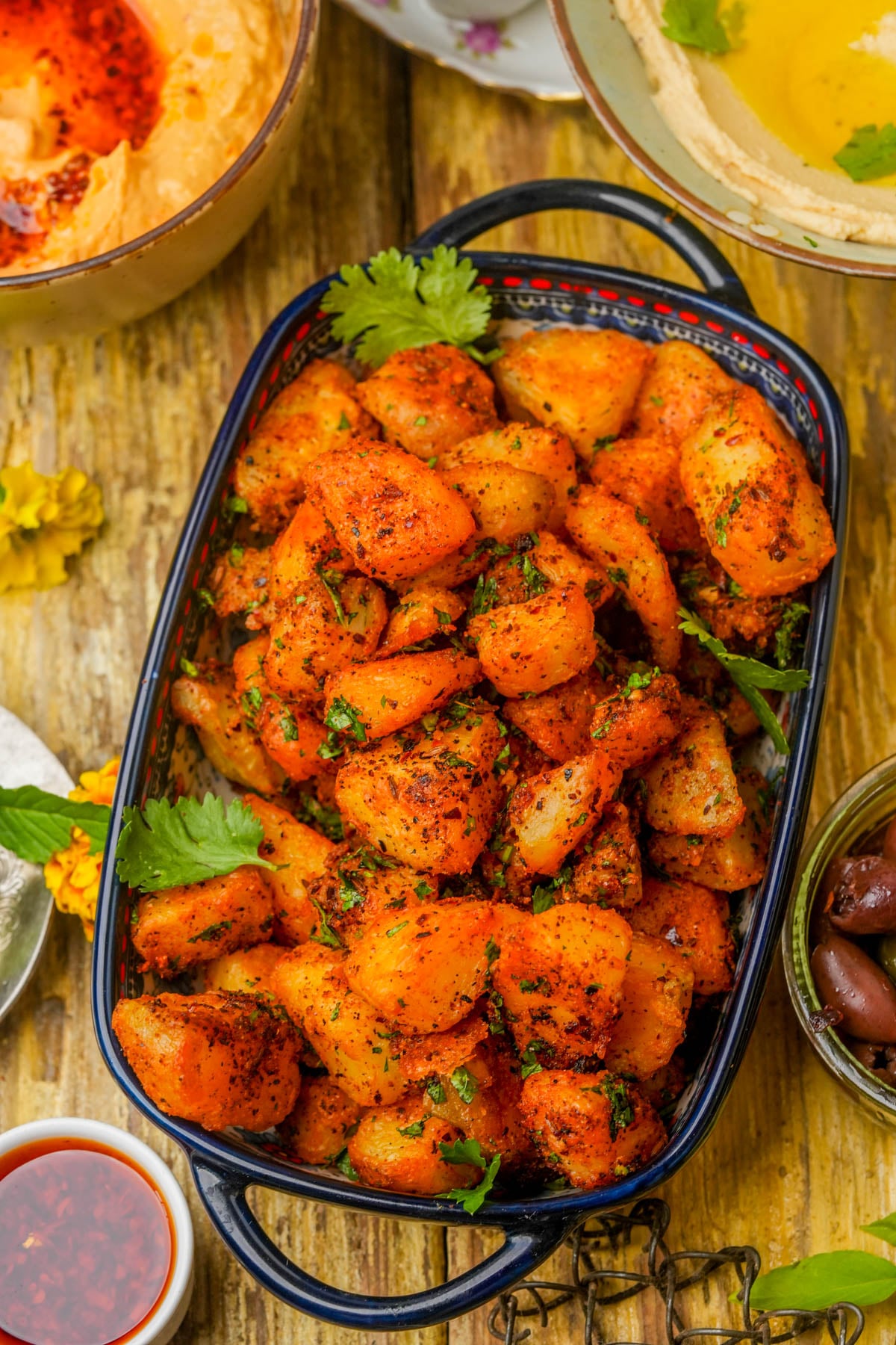 A black dish filled with seasoned, roasted batata harra garnished with fresh herbs, surrounded by small bowls of hummus, olives, and sauce on a rustic wooden table.