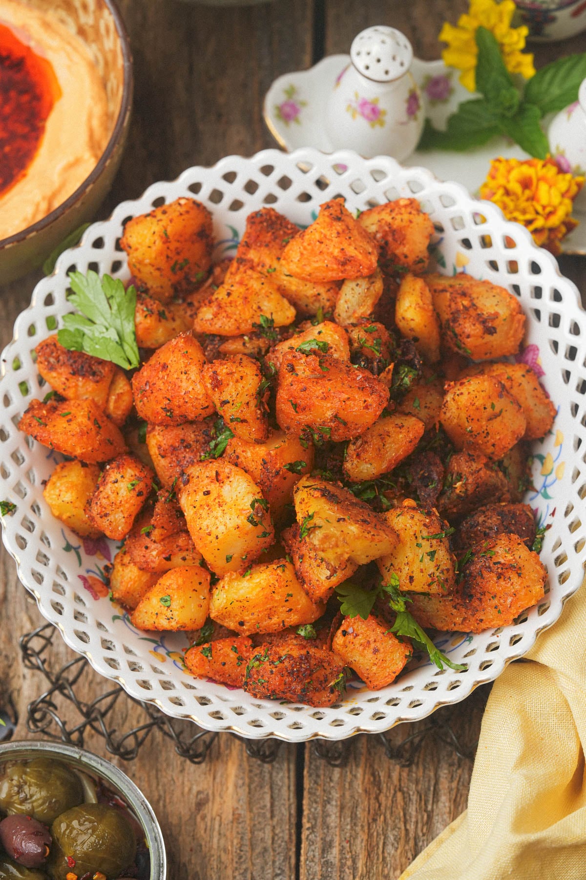 A white lattice bowl filled with seasoned, batata harra garnished with fresh herbs, placed on a rustic wooden table with olives, hummus, and decorative flowers nearby.