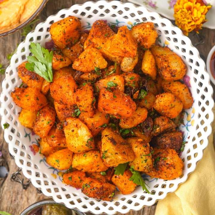 A white basket-weave bowl filled with golden, crispy batata harra garnished with parsley, sitting on a rustic wooden table.