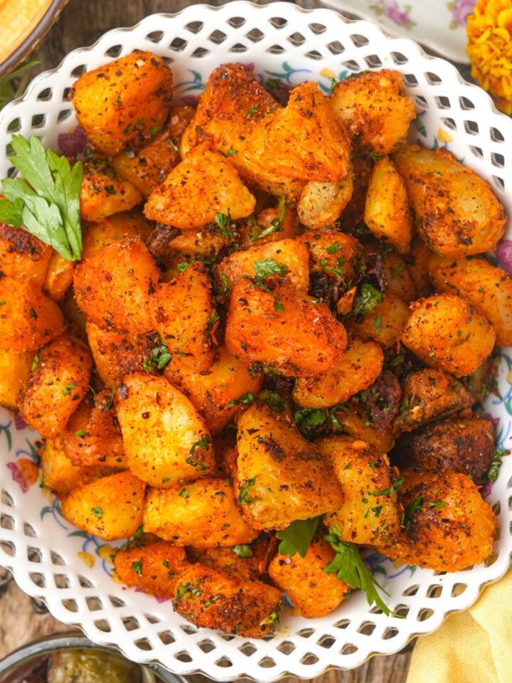 A white basket-weave bowl filled with golden, crispy batata harra garnished with parsley, sitting on a rustic wooden table.