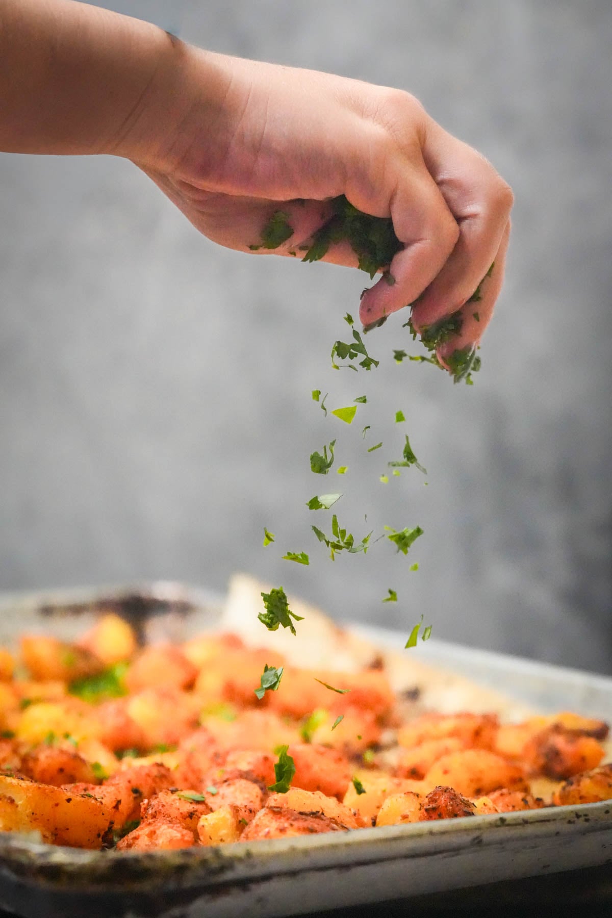 A hand sprinkles fresh chopped herbs over a tray of roasted batata harra.