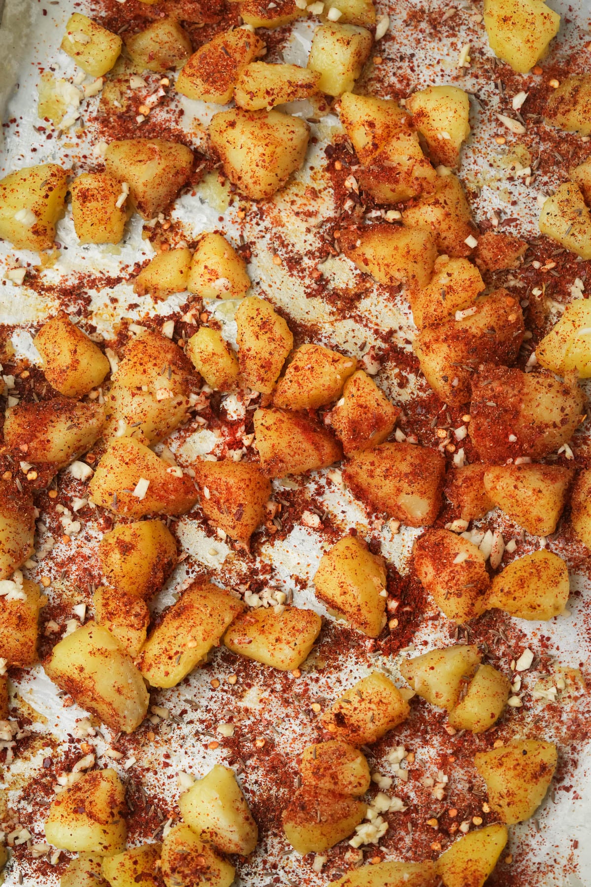 Chunks of potatoes coated with red spices and small pieces of garlic are spread out on a baking sheet lined with parchment paper, ready for roasting.