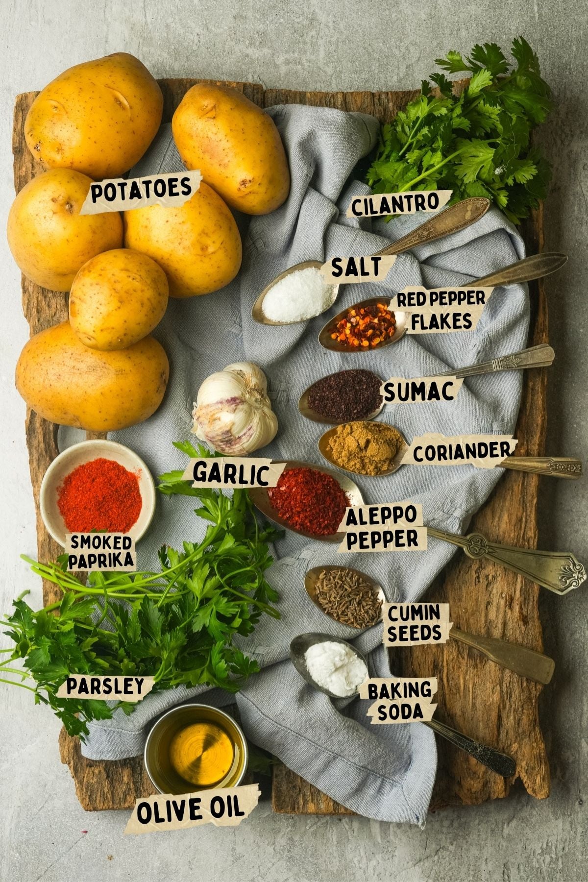 Five potatoes, fresh parsley, cilantro, garlic, a small bowl of olive oil, and spoons holding salt, sumac, red pepper flakes, smoked paprika, Aleppo pepper, coriander, cumin seeds, and baking soda on a wooden board.