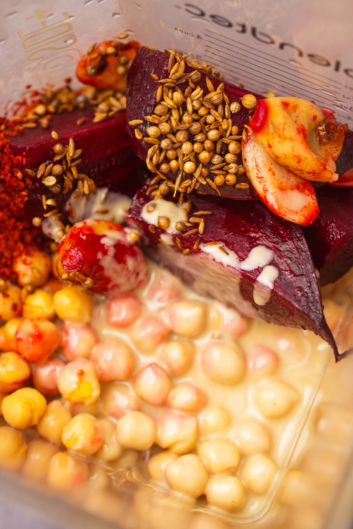 Chickpeas, beetroot, garlic, coriander seeds, chili flakes, and tahini in a blender, ready to be blended for a beet hummus recipe.