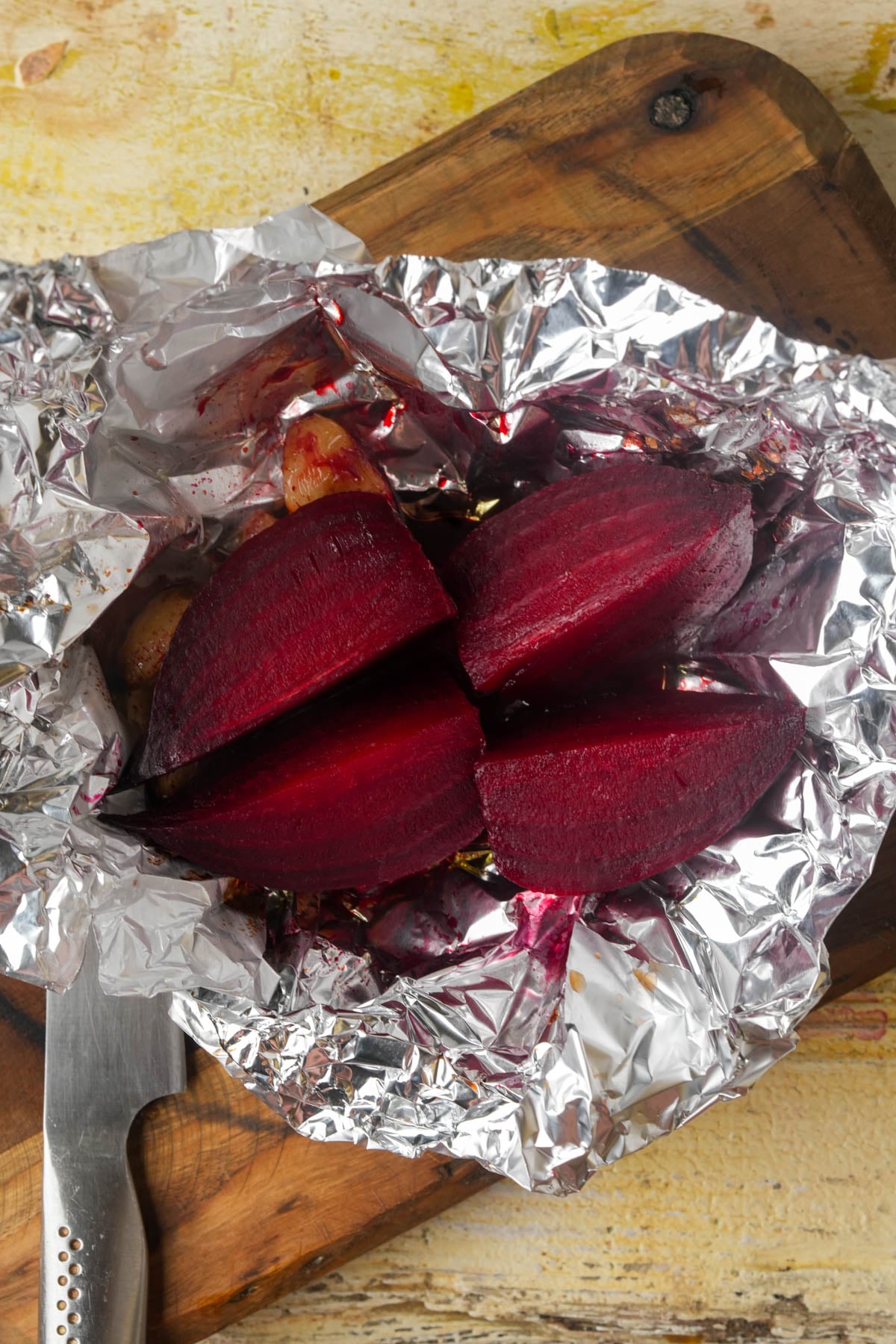 Roasted beetroot cut into quarters sits on crumpled aluminum foil atop a wooden cutting board, with a knife placed nearby.